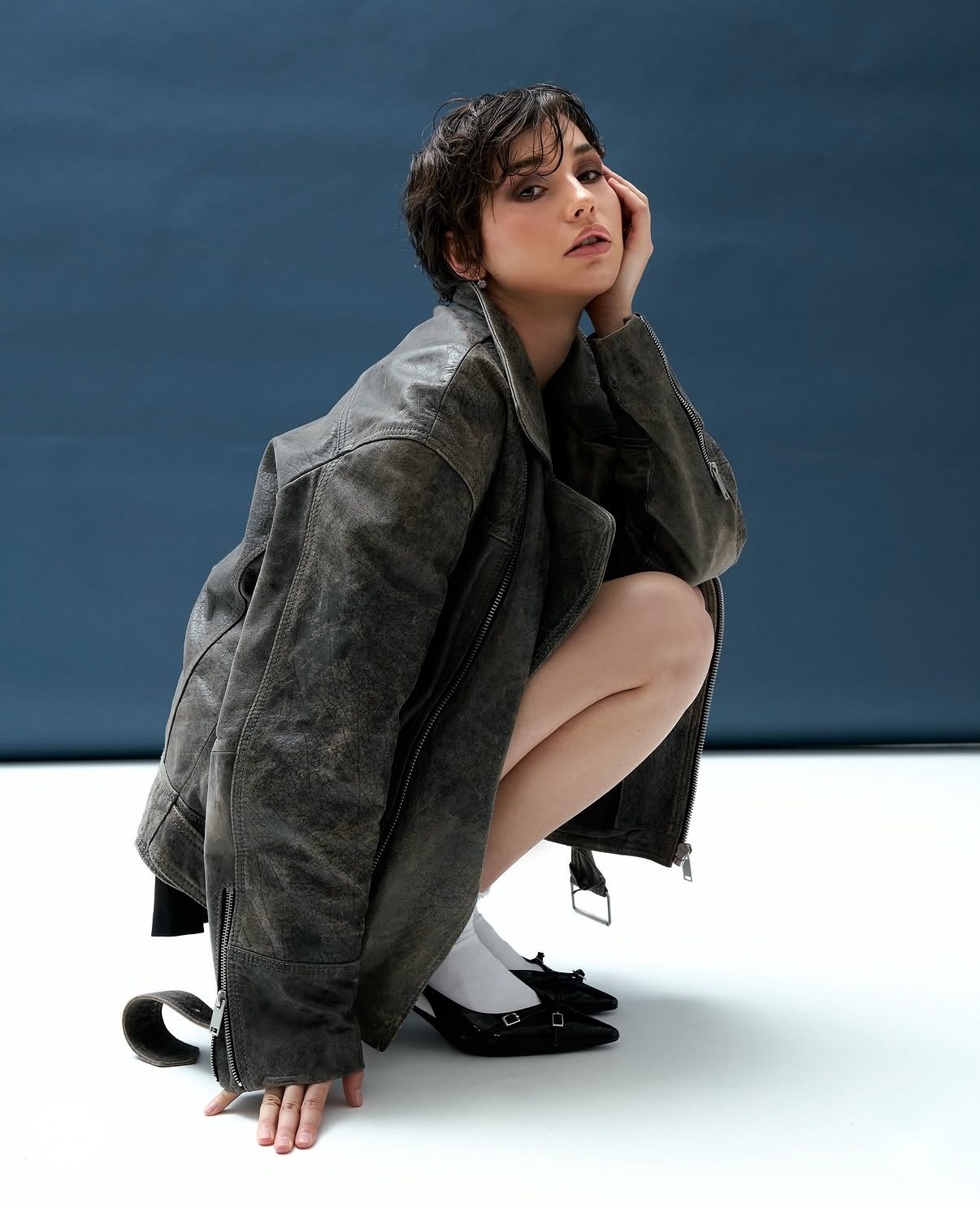 Model with a pixie cut crouching in a distressed leather jacket on a blue backdrop, shot by a previous workshop participant