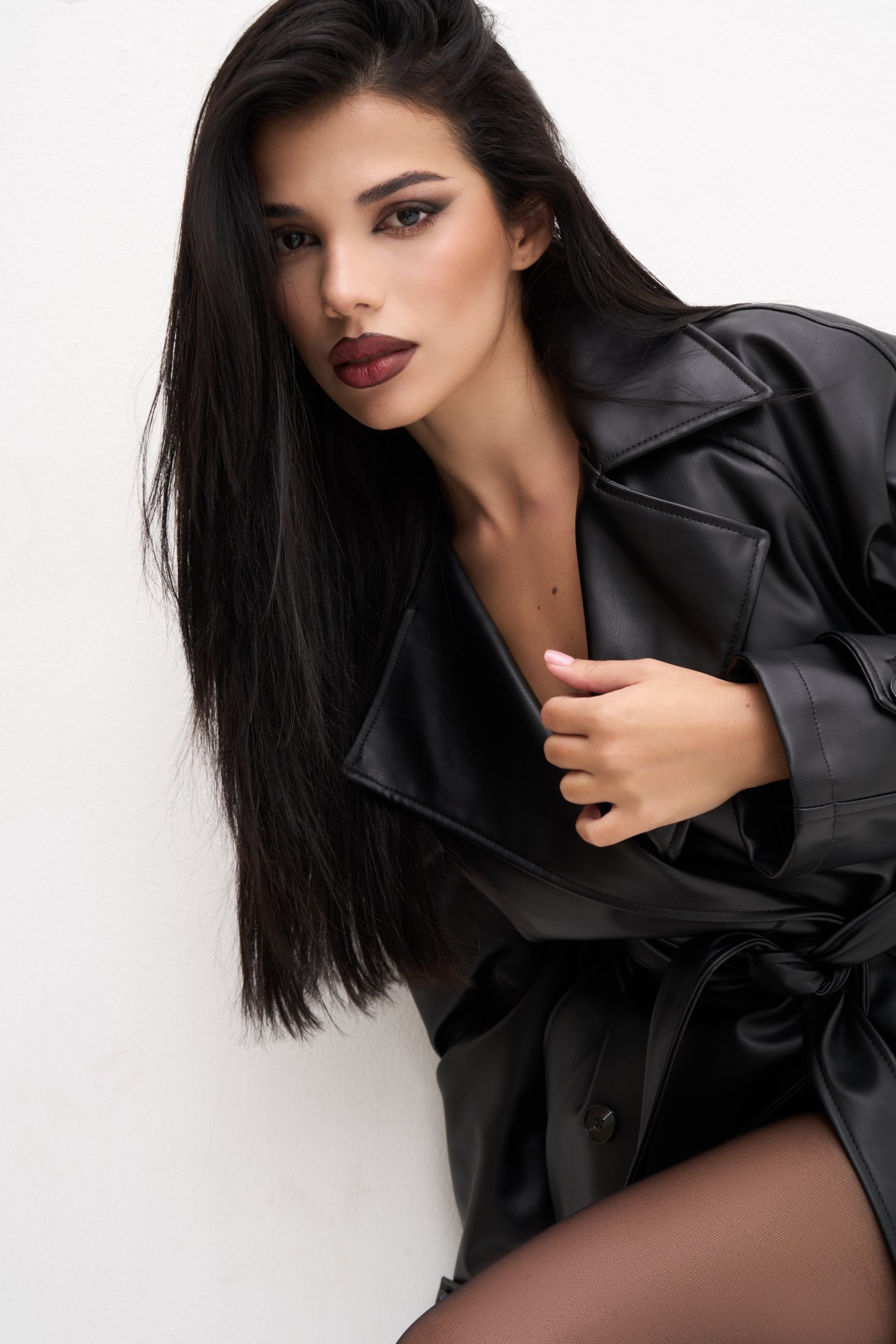 Studio portrait in a black leather trench against a white wall